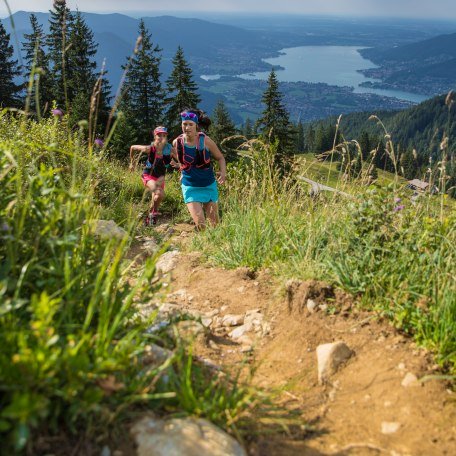 Ein schmaler Trail führt den Berg hinauf im Hintergrund sieht man zwei Läuferinnen und den Tegernsee., © Hansi Heckmair Ein schmaler Trail führt den Berg hinauf im Hintergrund sieht man zwei Läuferinnen und den Tegernsee., © Hansi Heckmair
