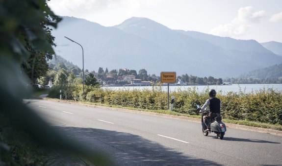 Rollerfahrer passiert die Ortseinfahrt der Stadt Tegernsee, im Hintergrund sieht man den Tegernsee., &copy; Der Tegernsee, Hansi Heckmair