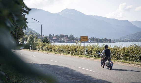 Rollerfahrer passiert die Ortseinfahrt der Stadt Tegernsee, im Hintergrund sieht man den Tegernsee., © Der Tegernsee, Hansi Heckmair Rollerfahrer passiert die Ortseinfahrt der Stadt Tegernsee, im Hintergrund sieht man den Tegernsee., © Der Tegernsee, Hansi Heckmair