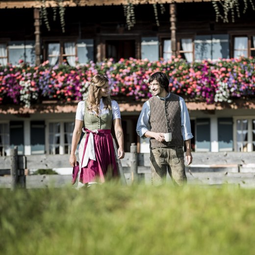 Pärchen in Tracht, schlendert über Blumenwiese vor altem mit Geranien geschmücktem Bauernhaus., © Hansi Heckmair